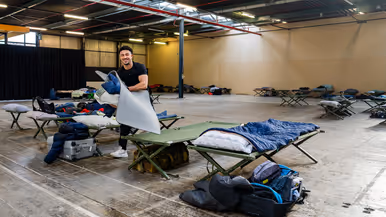 Van links naar rechts: een jongeman pakt iets van een veldbedje met een slaapzaak in een grote zaal. Een militair in een blauw uniform legt iets uit aan een groepje toehoorders in een donkere zaal. Twee man zitten aan een schaakbord met een poster van Defensie op de achtergrond. Een man in uniform met strepen op de schouder in bespreking met vijf anderen.