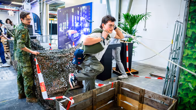 Diverse beelden van de fysieke uitdagingen, van links naar rechts: een team loopt met de handen aan een touw, waarbij ze over hindernissen moeten komen. Een team is gemaskerd en moet een ei in een dop over een parcours leiden. Rechts hangen twee deelnemers aan een rekstok.