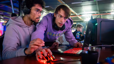Van links naar rechts: in een zaal met veel tafels sluit een jongen zijn apparatuur aan op een computer, die lichtgevend op het bureau staat. Een man lijkt aanwijzingen te geven aan een gamer achter de computer, dan geven ze elkaar een high five.