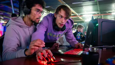 Van links naar rechts: in een zaal met veel tafels sluit een jongen zijn apparatuur aan op een computer, die lichtgevend op het bureau staat. Een man lijkt aanwijzingen te geven aan een gamer achter de computer, dan geven ze elkaar een high five.