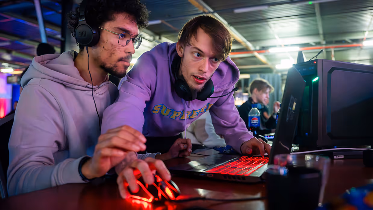 Van links naar rechts: in een zaal met veel tafels sluit een jongen zijn apparatuur aan op een computer, die lichtgevend op het bureau staat. Een man lijkt aanwijzingen te geven aan een gamer achter de computer, dan geven ze elkaar een high five.