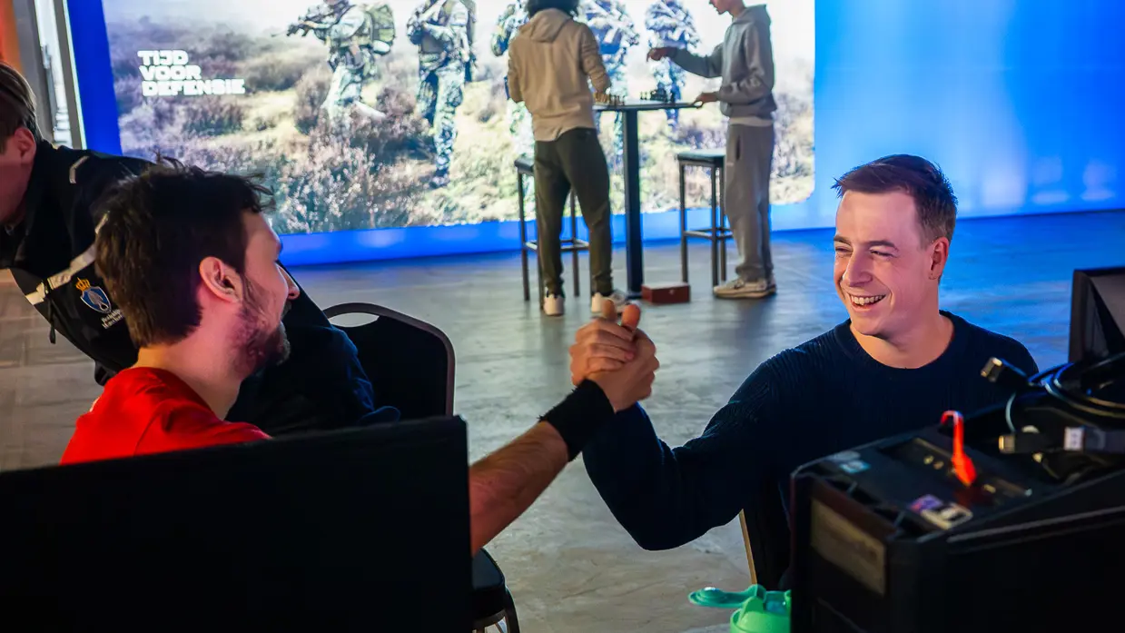 Van links naar rechts: in een zaal met veel tafels sluit een jongen zijn apparatuur aan op een computer, die lichtgevend op het bureau staat. Een man lijkt aanwijzingen te geven aan een gamer achter de computer, dan geven ze elkaar een high five.