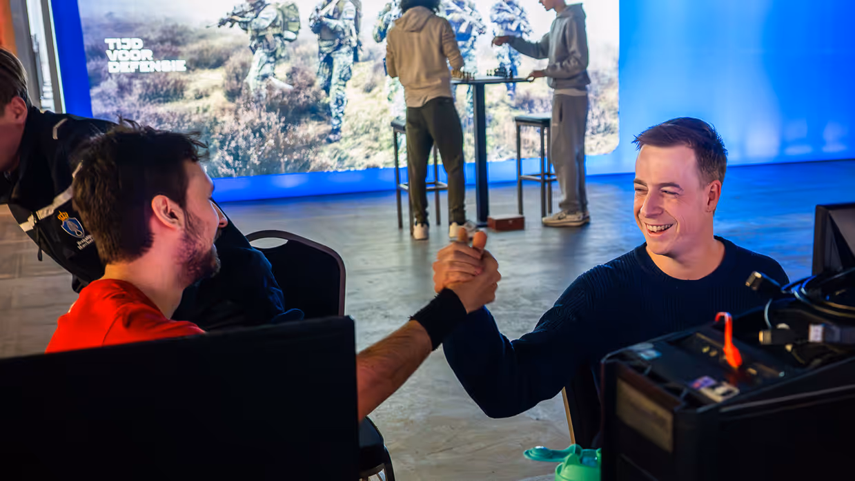 Van links naar rechts: in een zaal met veel tafels sluit een jongen zijn apparatuur aan op een computer, die lichtgevend op het bureau staat. Een man lijkt aanwijzingen te geven aan een gamer achter de computer, dan geven ze elkaar een high five.