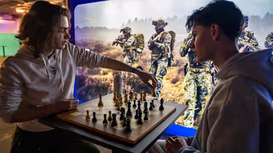 Van links naar rechts: een jongeman pakt iets van een veldbedje met een slaapzaak in een grote zaal. Een militair in een blauw uniform legt iets uit aan een groepje toehoorders in een donkere zaal. Twee man zitten aan een schaakbord met een poster van Defensie op de achtergrond. Een man in uniform met strepen op de schouder in bespreking met vijf anderen.