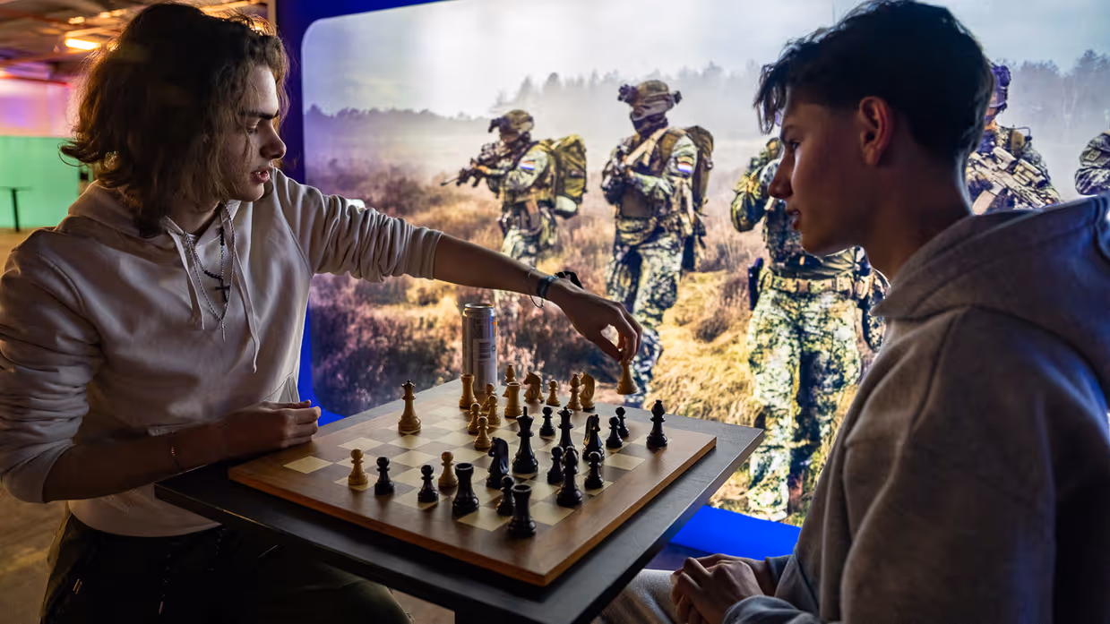 Van links naar rechts: een jongeman pakt iets van een veldbedje met een slaapzaak in een grote zaal. Een militair in een blauw uniform legt iets uit aan een groepje toehoorders in een donkere zaal. Twee man zitten aan een schaakbord met een poster van Defensie op de achtergrond. Een man in uniform met strepen op de schouder in bespreking met vijf anderen.