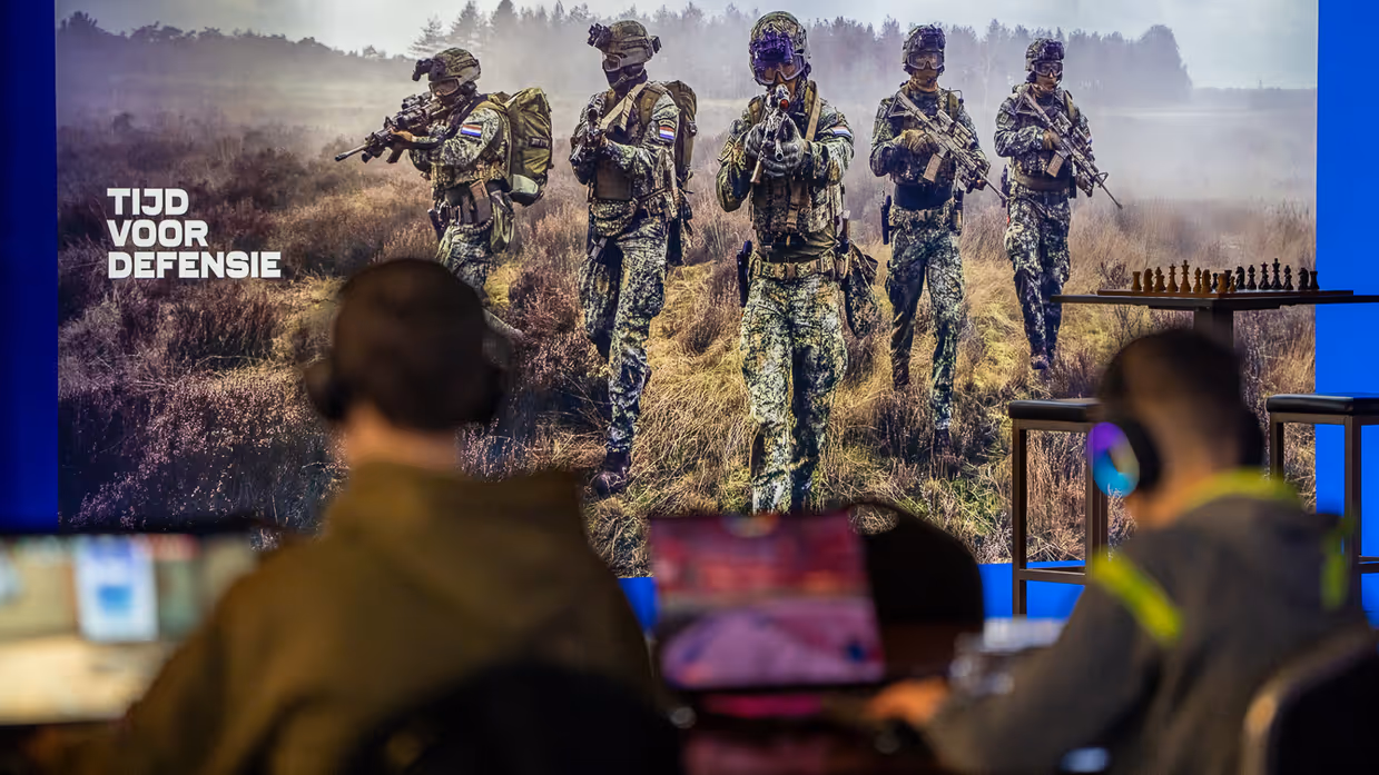 Met een poster van Tijd voor Defensie zitten twee man met computers vaag op de voorgrond.