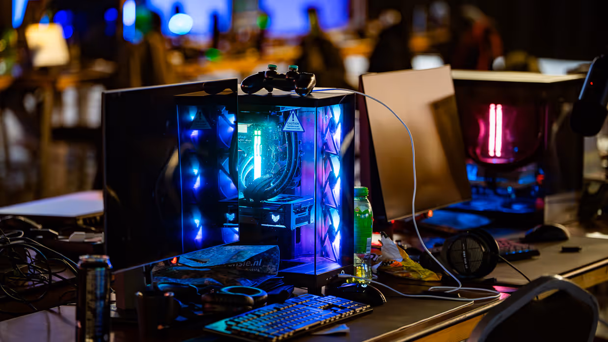 Van links naar rechts: in een zaal met veel tafels sluit een jongen zijn apparatuur aan op een computer, die lichtgevend op het bureau staat. Een man lijkt aanwijzingen te geven aan een gamer achter de computer, dan geven ze elkaar een high five.