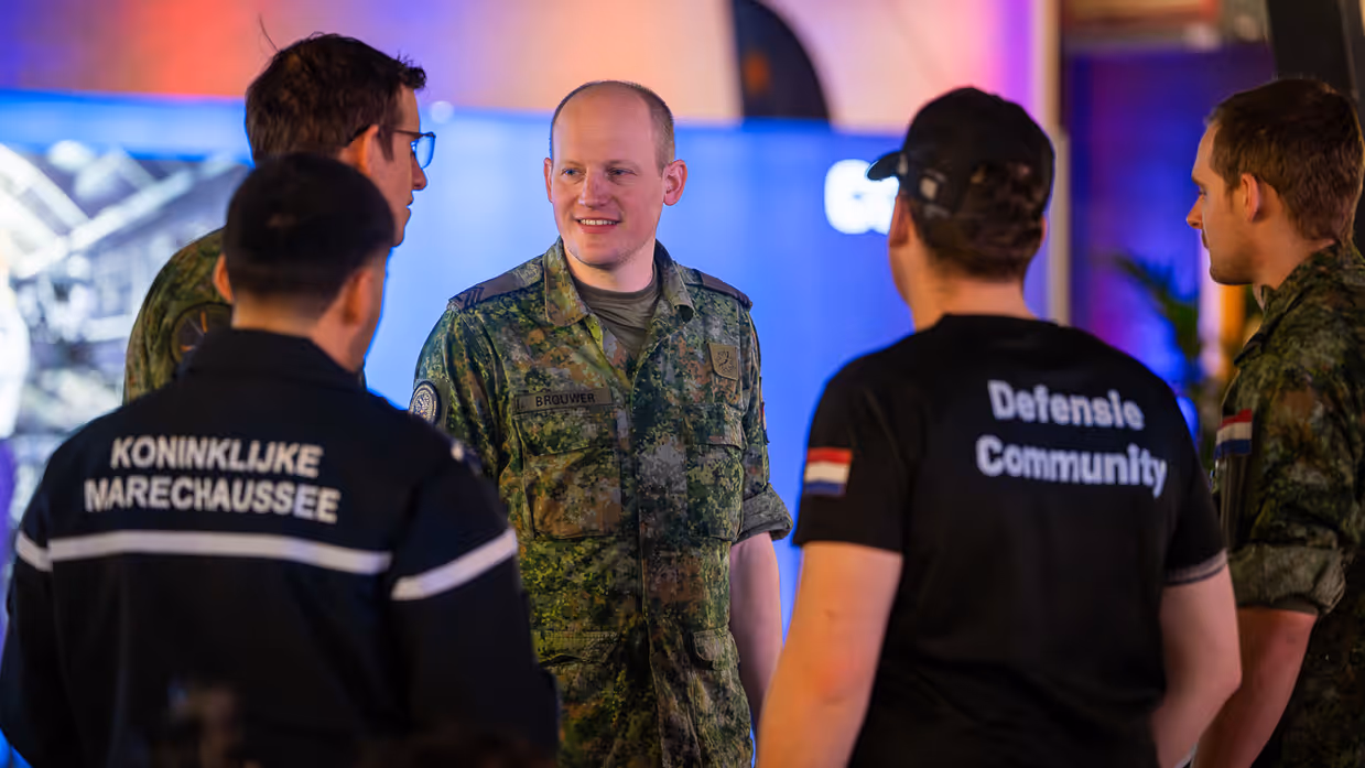 Van links naar rechts: een jongeman pakt iets van een veldbedje met een slaapzaak in een grote zaal. Een militair in een blauw uniform legt iets uit aan een groepje toehoorders in een donkere zaal. Twee man zitten aan een schaakbord met een poster van Defensie op de achtergrond. Een man in uniform met strepen op de schouder in bespreking met vijf anderen.