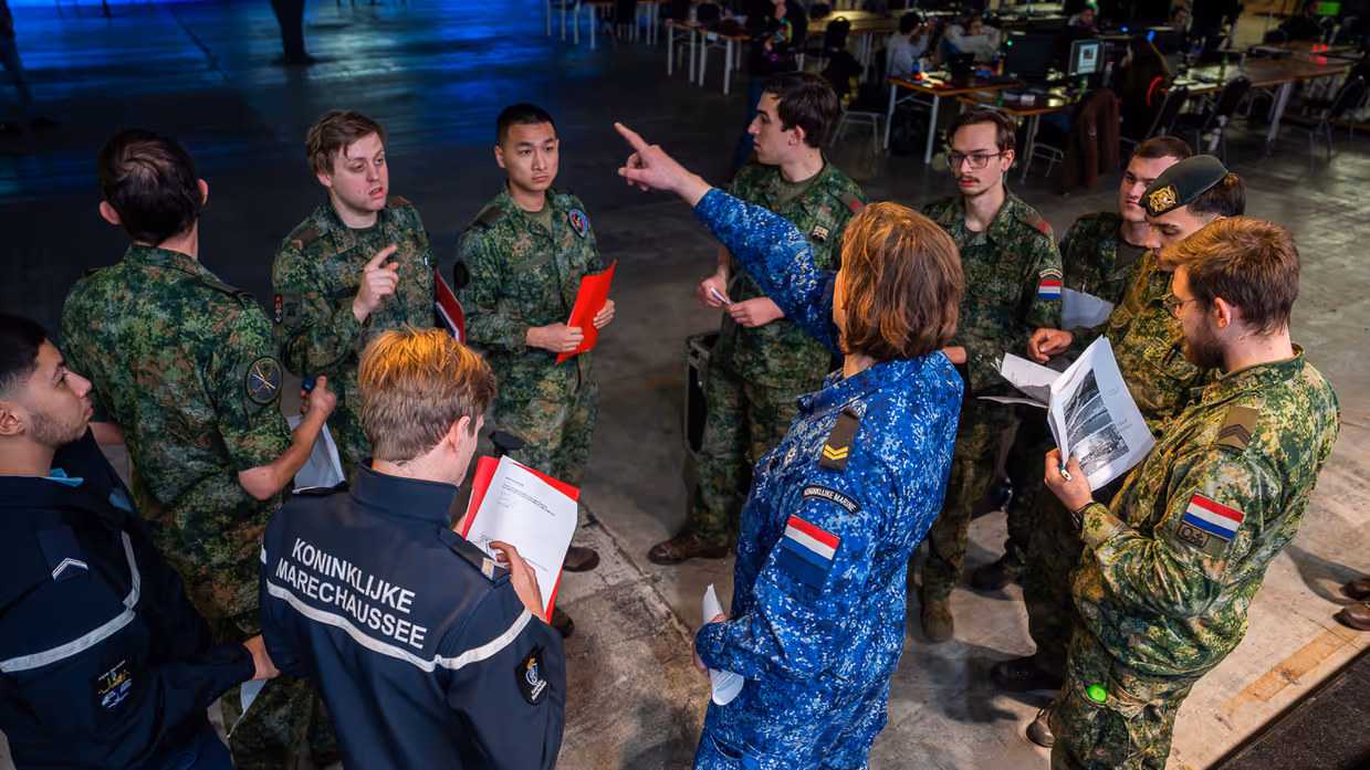 Van links naar rechts: een jongeman pakt iets van een veldbedje met een slaapzaak in een grote zaal. Een militair in een blauw uniform legt iets uit aan een groepje toehoorders in een donkere zaal. Twee man zitten aan een schaakbord met een poster van Defensie op de achtergrond. Een man in uniform met strepen op de schouder in bespreking met vijf anderen.