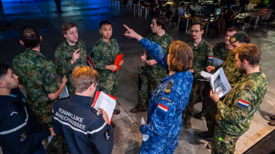 Van links naar rechts: een jongeman pakt iets van een veldbedje met een slaapzaak in een grote zaal. Een militair in een blauw uniform legt iets uit aan een groepje toehoorders in een donkere zaal. Twee man zitten aan een schaakbord met een poster van Defensie op de achtergrond. Een man in uniform met strepen op de schouder in bespreking met vijf anderen.