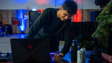 Van links naar rechts: in een zaal met veel tafels sluit een jongen zijn apparatuur aan op een computer, die lichtgevend op het bureau staat. Een man lijkt aanwijzingen te geven aan een gamer achter de computer, dan geven ze elkaar een high five.