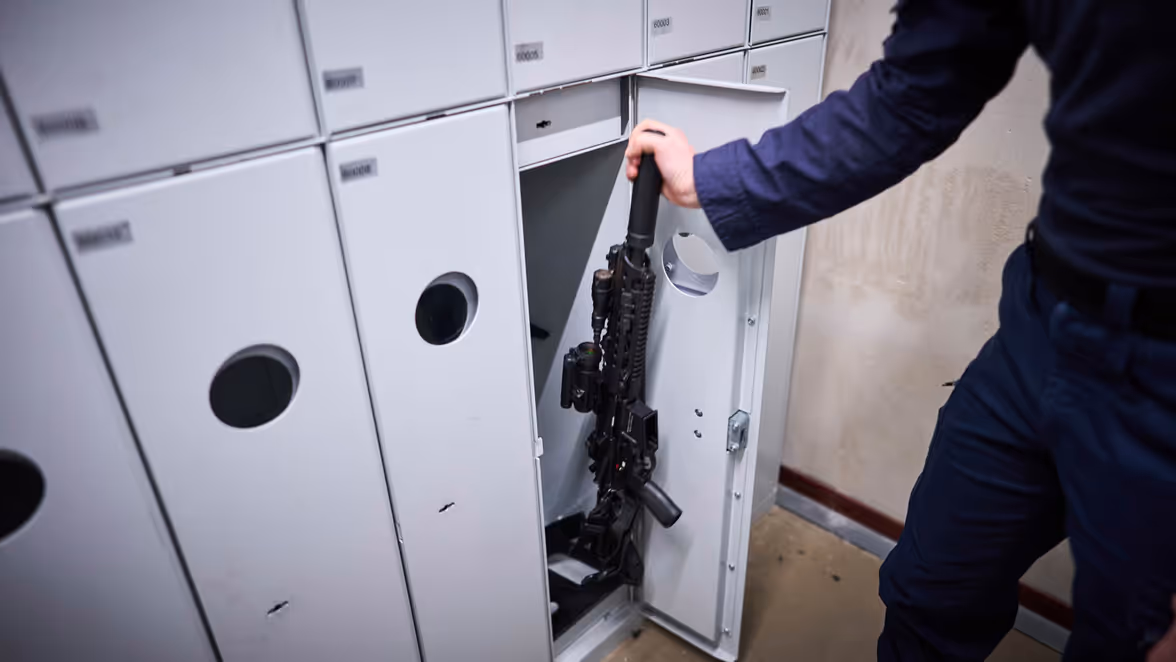 Een marechaussee-collega in uniform pakt zijn lange wapen uit een kluis