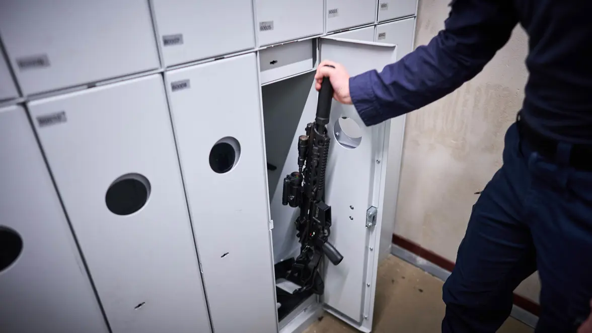 Een marechaussee-collega in uniform pakt zijn lange wapen uit een kluis