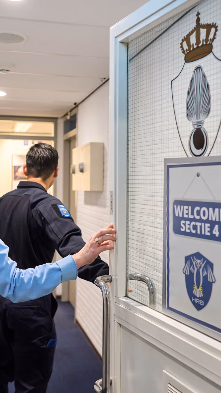 Adjudant Carolien en kapitein Tobias stappen door een deur waarop ‘Welcome Sectie 4’ wordt afgebeeld.