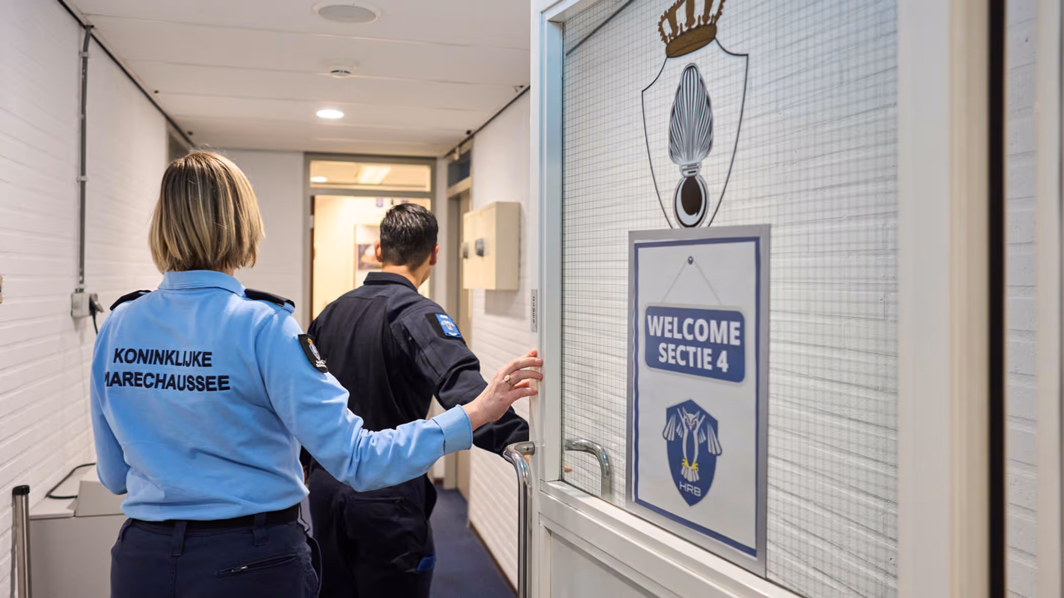 Adjudant Carolien en kapitein Tobias stappen door een deur waarop ‘Welcome Sectie 4’ wordt afgebeeld.