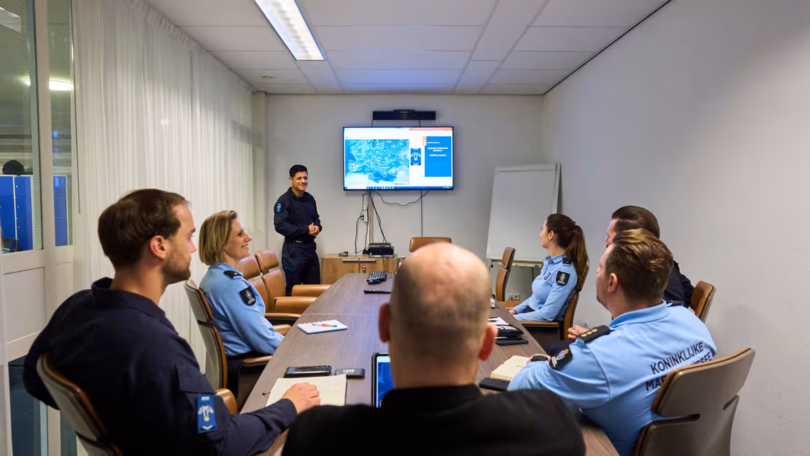 Marechausseecollega’s in uniform zitten aan een ovale tafel en kijken naar een digibord waarop een wereldkaart staat afgebeeld. 