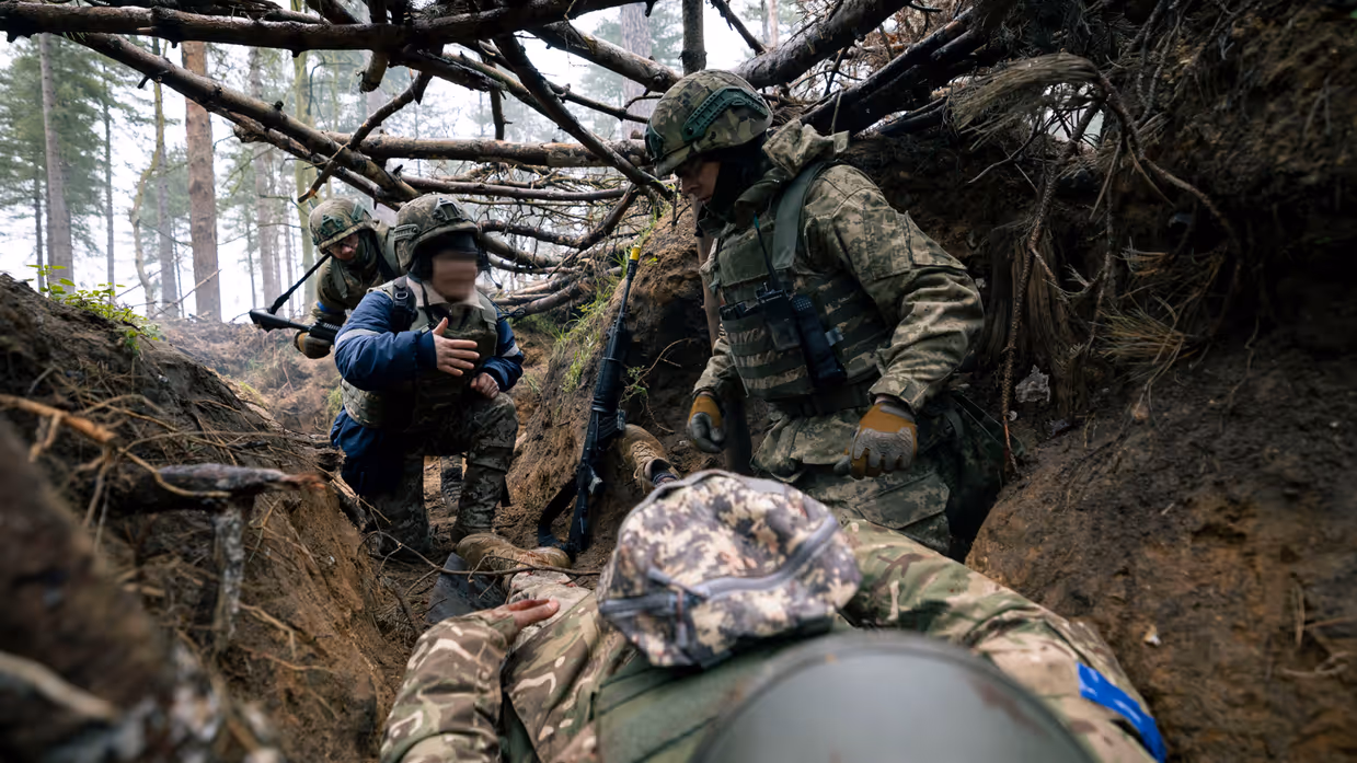 Drie Oekraïense militairen in een loopgraaf. Een tolk geeft instructies door bij een casus Zelfhulp Kameradenhulp (ZHKH). Een van de militairen neemt hierbij de rol aan van gewonde.