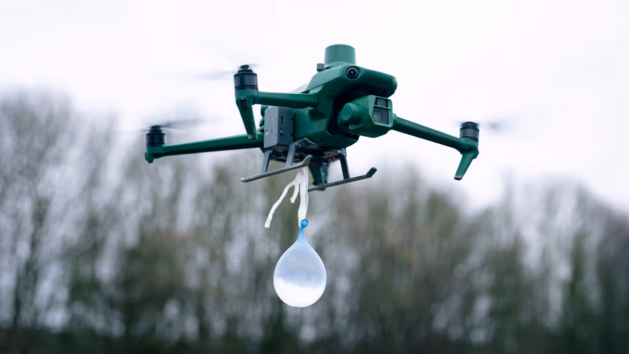 Een drone hangt laag in de lucht met een waterballon eronder.