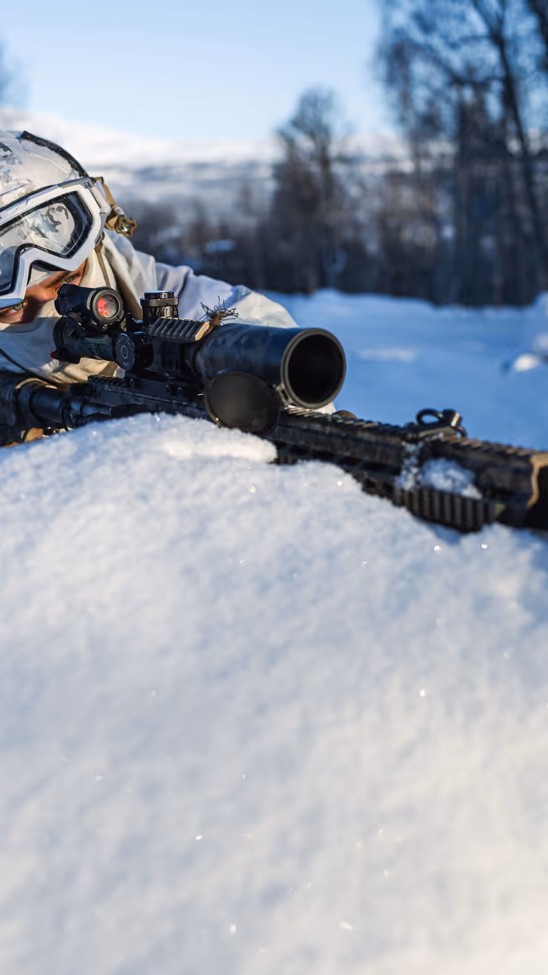 Militair in wintercamouflage ligt in de sneeuw en richt met een geweer tijdens een oefening in een besneeuwd landschap.