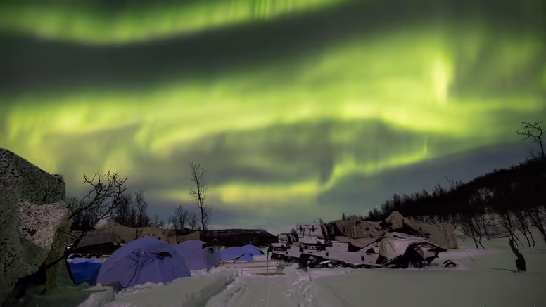 Een tentenkamp met sneeuwscooters in een besneeuwd landschap, onder het Noorderlicht.