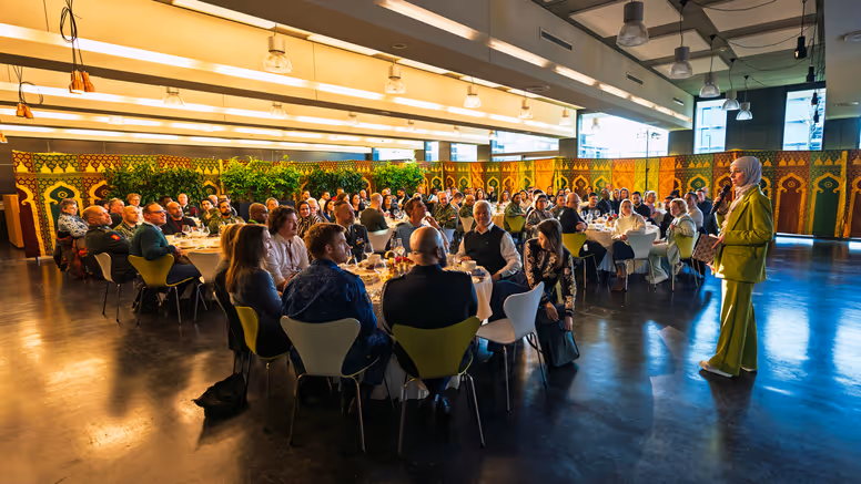 Een vrouw in groen pak staat voorin een ruimte en spreekt met microfoon, terwijl veel mensen aan ronde tafels zitten te luisteren en eten. 