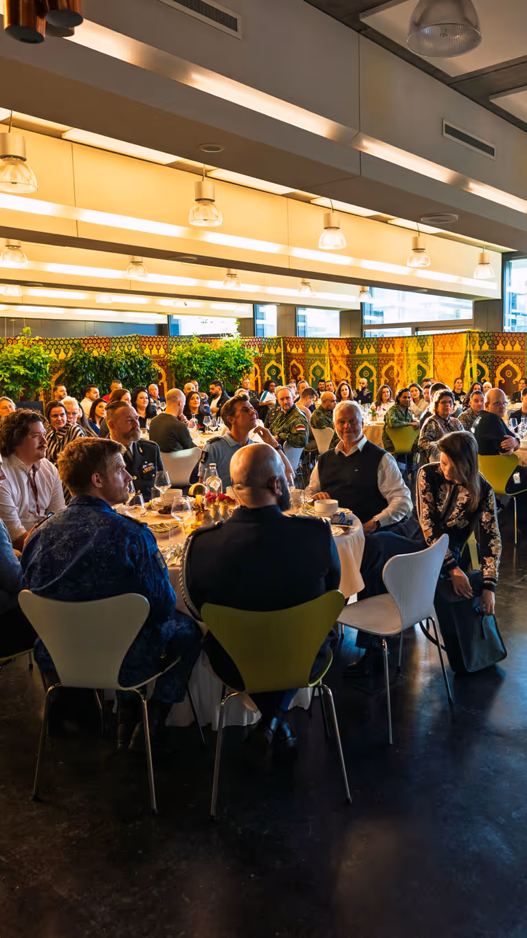 Een vrouw in groen pak staat voorin een ruimte en spreekt met microfoon, terwijl veel mensen aan ronde tafels zitten te luisteren en eten. 