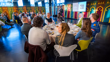  Links: Een grote, lichte ruimte waar mensen aan ronde tafels zitten te eten. Vooraan staat een man in uniform te spreken, terwijl de rest luistert. Rechts: Een glimlachende man in militair uniform met medailles spreekt in een microfoon, terwijl een andere man in uniform naar hem luistert met een boeket bloemen op tafel.