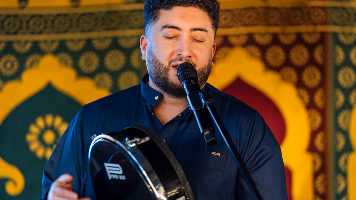 Links: Een man in een donkerblauw uniform met gouden versieringen staat voor een groep mensen en spreekt, terwijl het publiek naar hem luistert. Rechts een man met gesloten ogen die zingt in een microfoon. Hij houdt een handtrommel vast en draagt een donkerblauwe kleding, omringd door kleurrijke patronen.