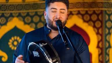 Links: Een man in een donkerblauw uniform met gouden versieringen staat voor een groep mensen en spreekt, terwijl het publiek naar hem luistert. Rechts een man met gesloten ogen die zingt in een microfoon. Hij houdt een handtrommel vast en draagt een donkerblauwe kleding, omringd door kleurrijke patronen.