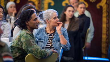 Twee mensen zitten naast elkaar in een zaal: een vrouw met grijs haar en een denim jasje luistert aandachtig, een man met lang haar kijkt mee.