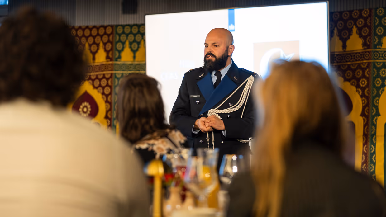 Links: Een man in een donkerblauw uniform met gouden versieringen staat voor een groep mensen en spreekt, terwijl het publiek naar hem luistert. Rechts een man met gesloten ogen die zingt in een microfoon. Hij houdt een handtrommel vast en draagt een donkerblauwe kleding, omringd door kleurrijke patronen.