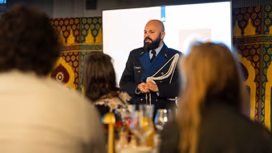 Links: Een man in een donkerblauw uniform met gouden versieringen staat voor een groep mensen en spreekt, terwijl het publiek naar hem luistert. Rechts een man met gesloten ogen die zingt in een microfoon. Hij houdt een handtrommel vast en draagt een donkerblauwe kleding, omringd door kleurrijke patronen.