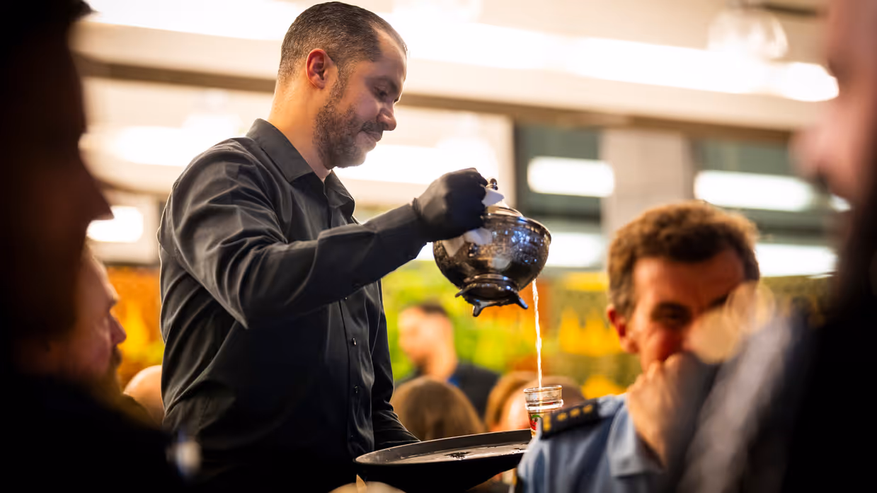 Links: Een man in zwart overhemd schenkt met een glimlach thee uit een traditionele theepot in een klein glas voor een gast, terwijl anderen toekijken. Rechts: Twee mensen zitten naast elkaar in een zaal: een vrouw met grijs haar en een denim jasje luistert aandachtig, een man met lang haar kijkt mee.