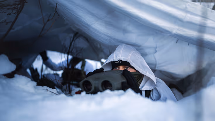 Een marinier met verrekijker onder wit zeildoek in de sneeuw.