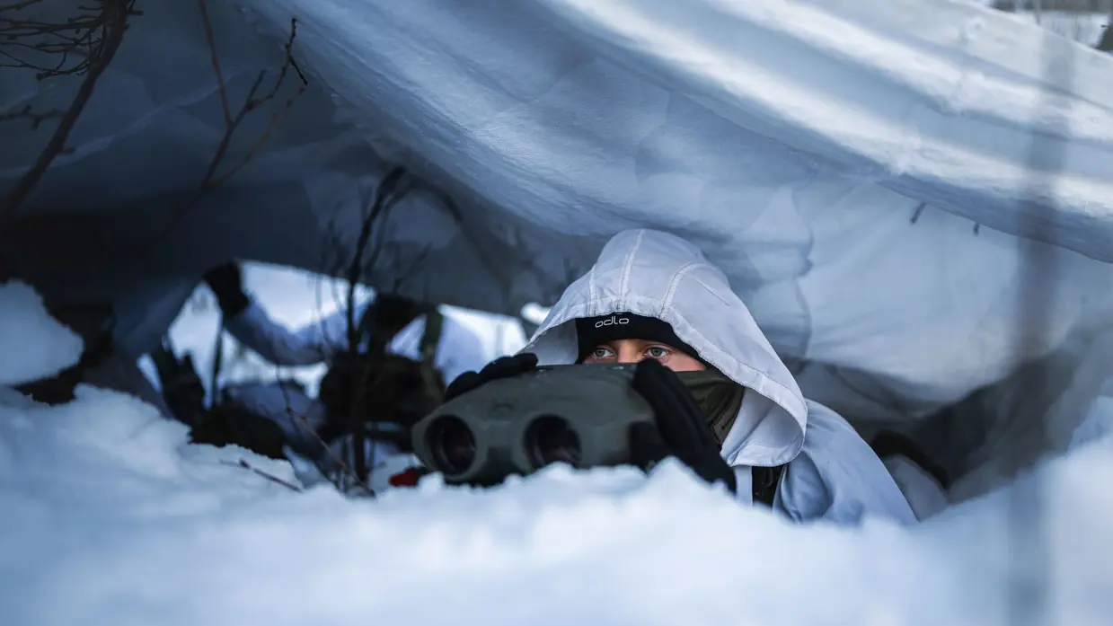 Een marinier met verrekijker onder wit zeildoek in de sneeuw.