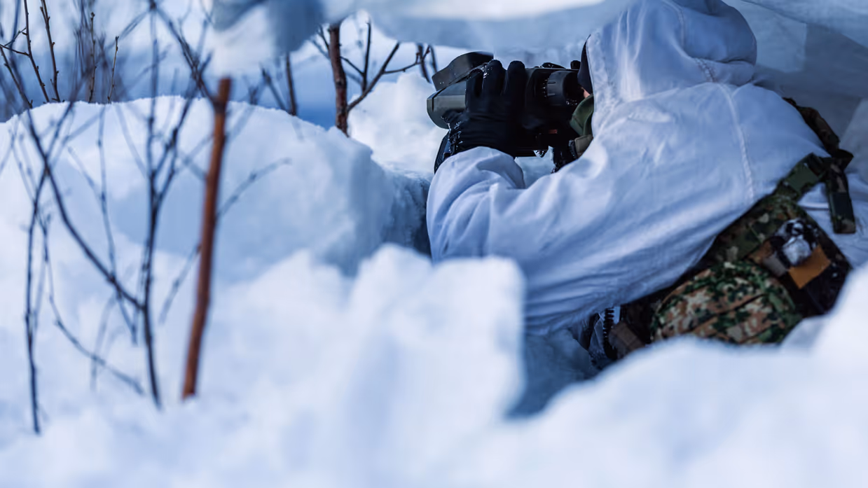 Militair in wintercamouflage observeert met een verrekijker vanuit een schuilpositie in de sneeuw.