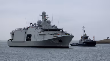 De Vlissingen met daarnaast een sleepboot, gezien vanaf de kade. Daarnaast het grijze marineschip van dichterbij, de sleepboot er nog altijd naast varend.