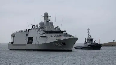 De Vlissingen met daarnaast een sleepboot, gezien vanaf de kade. Daarnaast het grijze marineschip van dichterbij, de sleepboot er nog altijd naast varend.