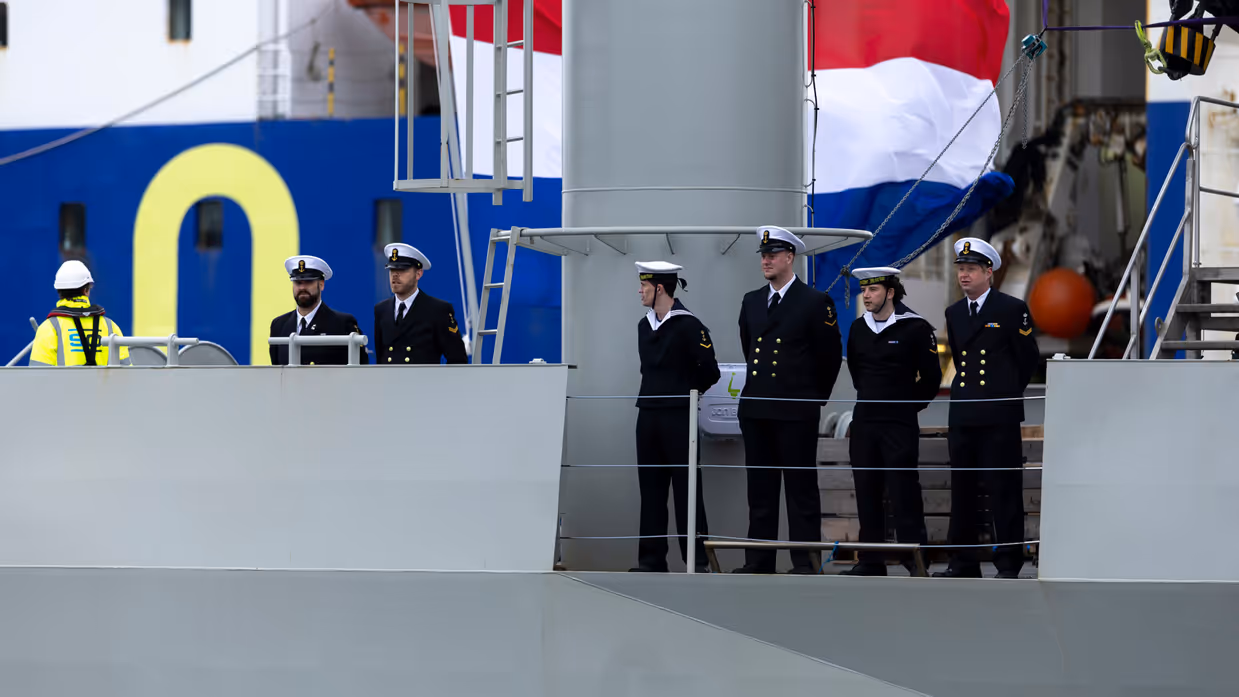 : Het varende, grijze marineschip vanaf de zijkant. Daarnaast een afbeelding met daarop een deel van de bemanning bij de reling van het schip, daarachter een wapperende Nederlandse vlag. Vervolgens saluerende militairen vanaf de brug van de Vlissingen en een groepje marineofficieren dat het schip vanaf de kade aanschouwt en vastlegt met de mobiele telefoon.