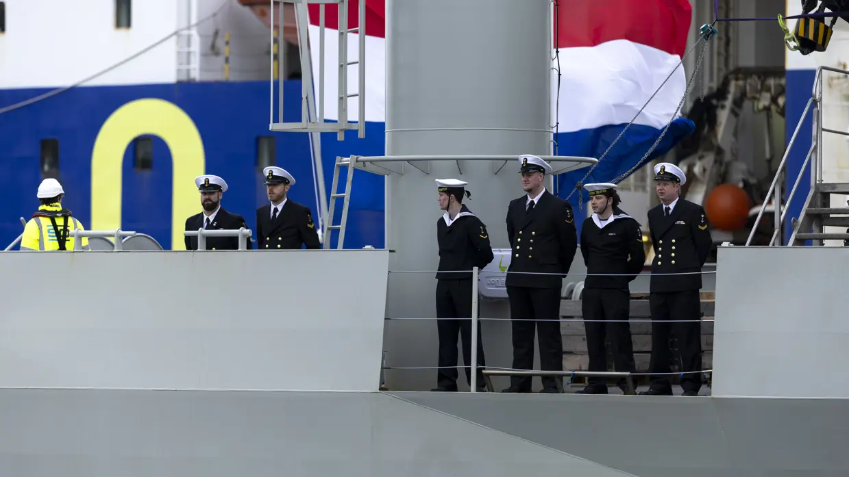 : Het varende, grijze marineschip vanaf de zijkant. Daarnaast een afbeelding met daarop een deel van de bemanning bij de reling van het schip, daarachter een wapperende Nederlandse vlag. Vervolgens saluerende militairen vanaf de brug van de Vlissingen en een groepje marineofficieren dat het schip vanaf de kade aanschouwt en vastlegt met de mobiele telefoon.