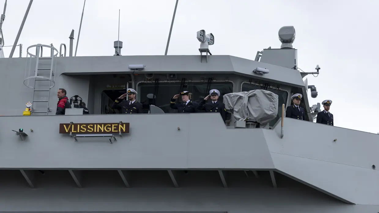 : Het varende, grijze marineschip vanaf de zijkant. Daarnaast een afbeelding met daarop een deel van de bemanning bij de reling van het schip, daarachter een wapperende Nederlandse vlag. Vervolgens saluerende militairen vanaf de brug van de Vlissingen en een groepje marineofficieren dat het schip vanaf de kade aanschouwt en vastlegt met de mobiele telefoon.