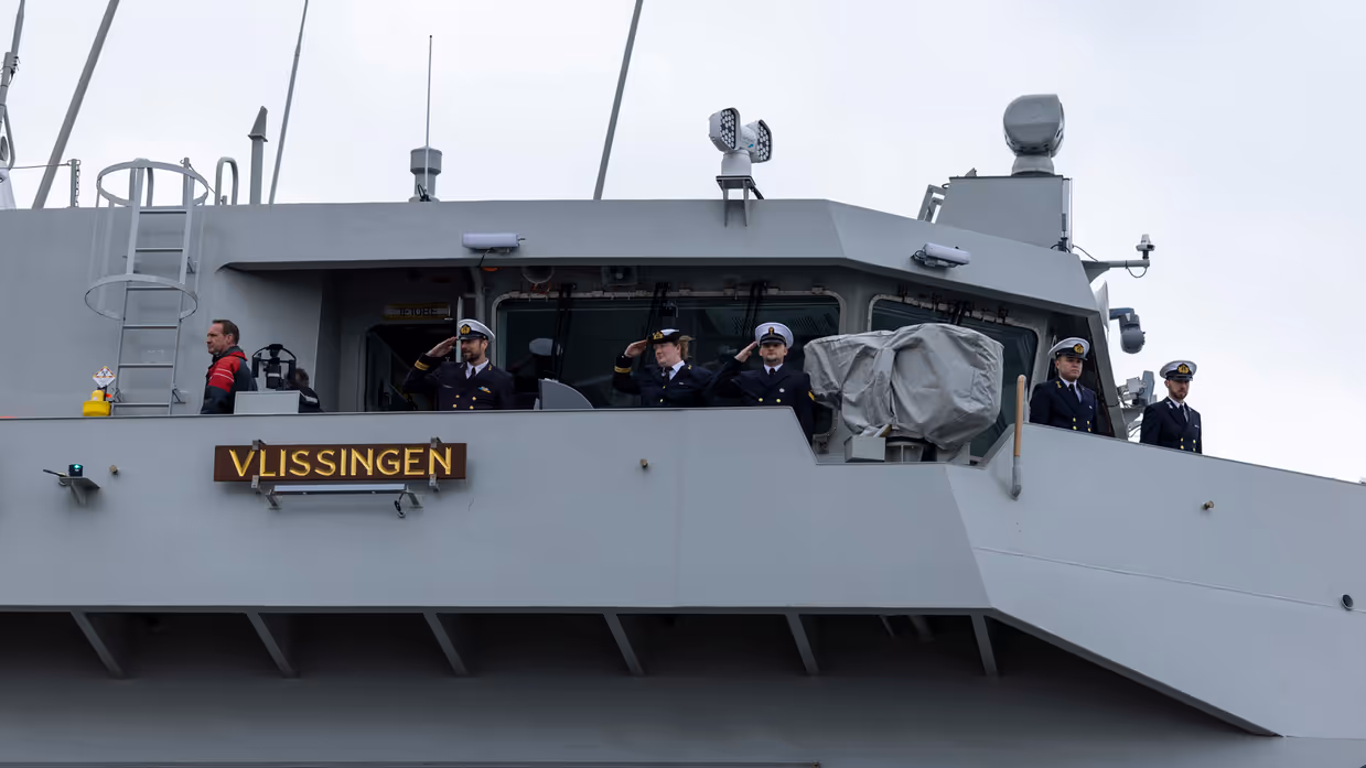 : Het varende, grijze marineschip vanaf de zijkant. Daarnaast een afbeelding met daarop een deel van de bemanning bij de reling van het schip, daarachter een wapperende Nederlandse vlag. Vervolgens saluerende militairen vanaf de brug van de Vlissingen en een groepje marineofficieren dat het schip vanaf de kade aanschouwt en vastlegt met de mobiele telefoon.