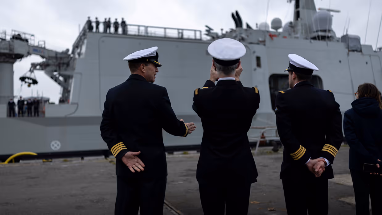 : Het varende, grijze marineschip vanaf de zijkant. Daarnaast een afbeelding met daarop een deel van de bemanning bij de reling van het schip, daarachter een wapperende Nederlandse vlag. Vervolgens saluerende militairen vanaf de brug van de Vlissingen en een groepje marineofficieren dat het schip vanaf de kade aanschouwt en vastlegt met de mobiele telefoon.