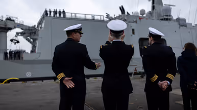 : Het varende, grijze marineschip vanaf de zijkant. Daarnaast een afbeelding met daarop een deel van de bemanning bij de reling van het schip, daarachter een wapperende Nederlandse vlag. Vervolgens saluerende militairen vanaf de brug van de Vlissingen en een groepje marineofficieren dat het schip vanaf de kade aanschouwt en vastlegt met de mobiele telefoon.