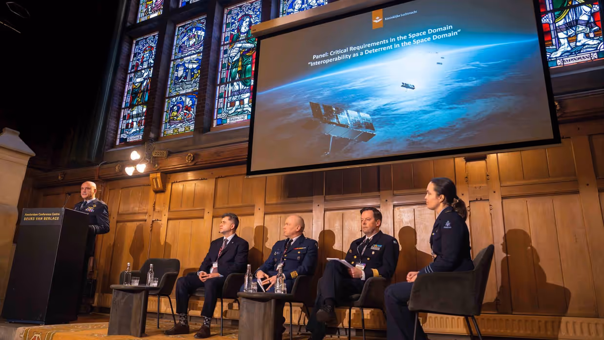 Vier mensen zitten op een podium met staand naast hen generaal Steur pratend vanachter een spreekgestoelte.