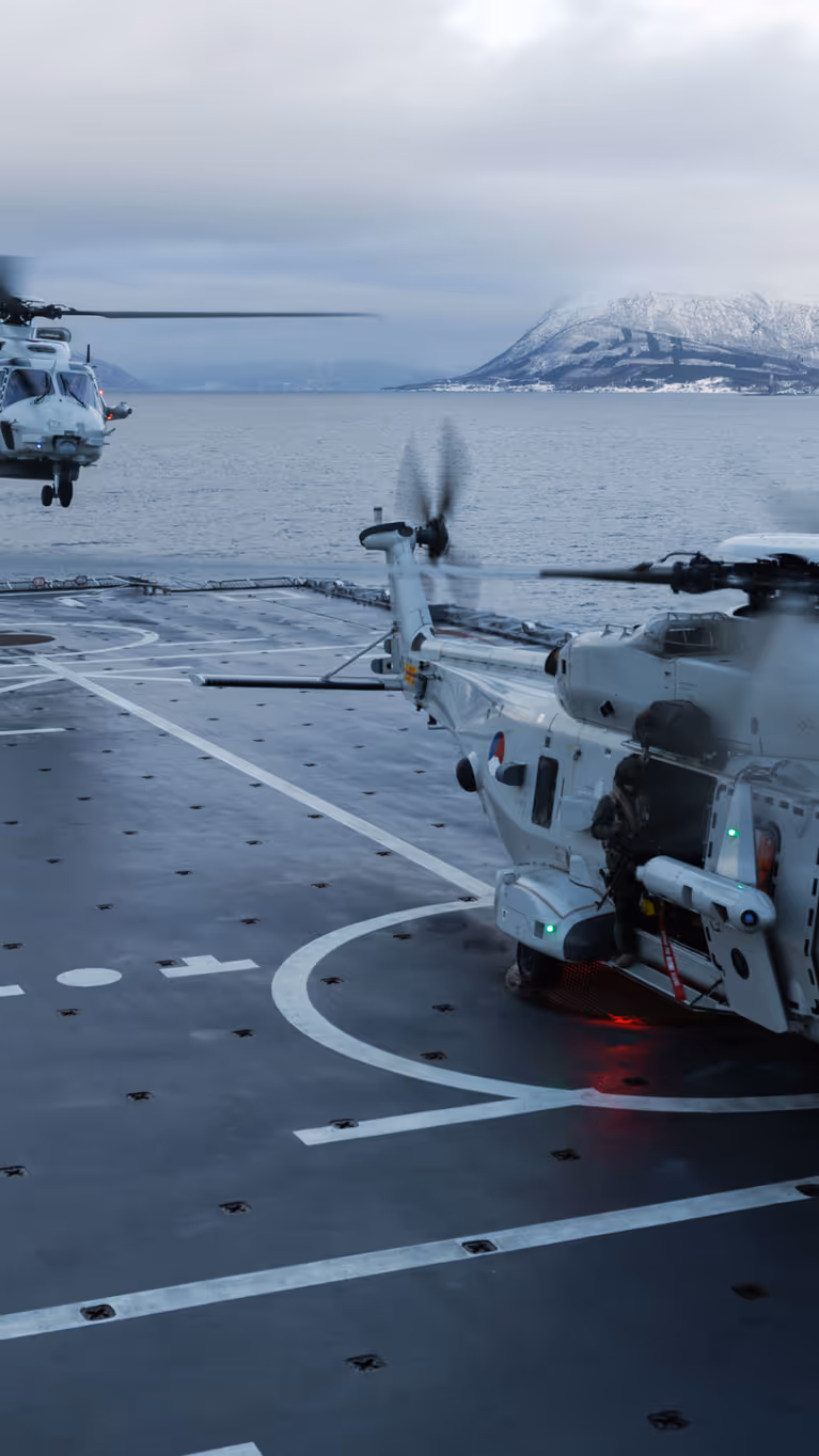 Twee NH90’s op het dek van Zr.Ms. Johan de Witt in een fjord met besneeuwde bergen; één staat op het dek terwijl de andere landt, en een vliegdekofficier geeft signalen.