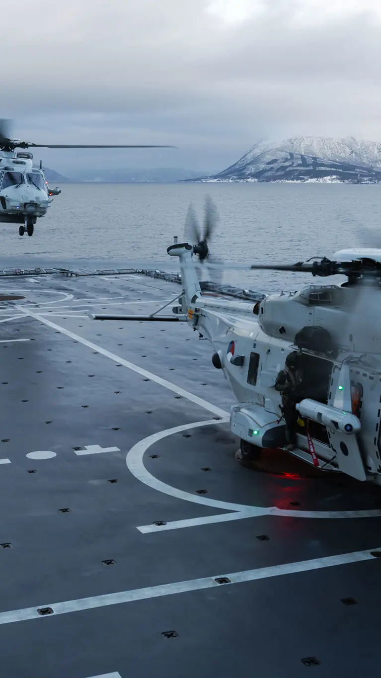Twee NH90’s op het dek van Zr.Ms. Johan de Witt in een fjord met besneeuwde bergen; één staat op het dek terwijl de andere landt, en een vliegdekofficier geeft signalen.