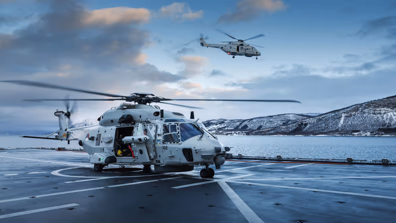 NH90-maritieme helikopter op het dek van een marineschip tijdens een oefening in een besneeuwd kustgebied, terwijl een tweede helikopter boven zee vliegt. 