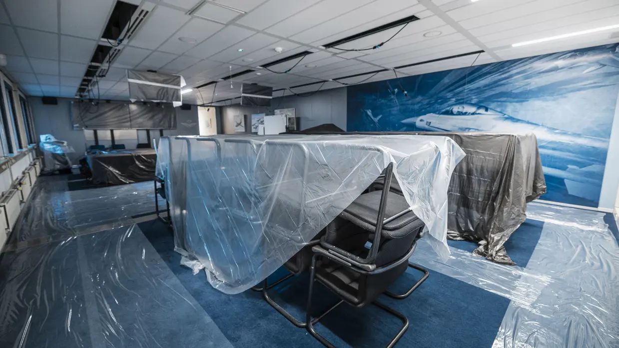 Vier foto’s van werkzaamheden. Naast een kantoorruimte met afgedekte bureaus, laptops en toetsenborden gaat het om een kabel die uit het plafond hangt. Tot slot het logo van de luchtmacht tegen een muur, daarnaast een afgedekt beeldscherm.