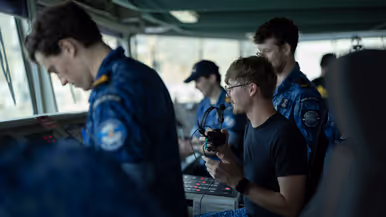 Links: Bemanningsleden bij de reling, met het scheepskanon op de achtergrond. Rechts: bemanning aan boord. Een van hen heeft een koptelefoon vast. 